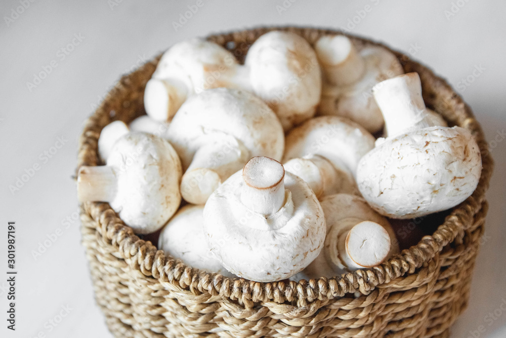 Mushrooms champignons in a round wicker basket on a white wooden table. Close up. Place for text or advertising Mushrooms champignons in a round wicker basket on a white wooden table. Close up. Place for text or advertising