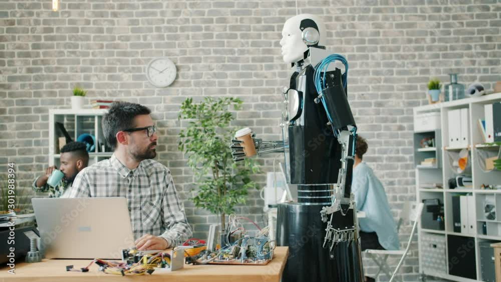 Slow motion of futuristic robot bringing coffee to young researcher in laboratory, guy is using compute then drinking from glass. Innovation and modern business concept.