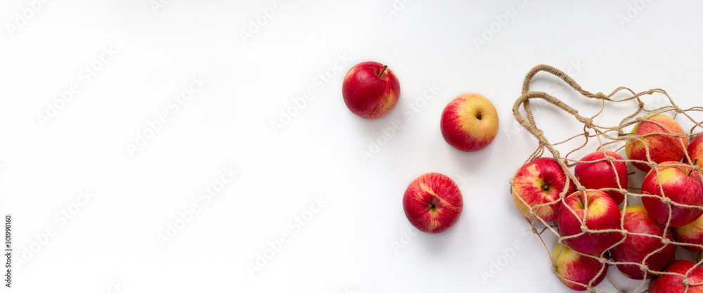 Fresh red apples in string bag on white background. Eco mesh with colorful fruits. Banner with copy space, top view, flat lay. Ecology and healthy eating concept