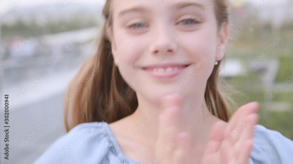 Charming long-haired 10 years old girl in a blue dress enthusiastically smiles and claps her hands, jumps, enjoys the good news, surprise. Slow motion, close-up. Cityscape in defocus on the background