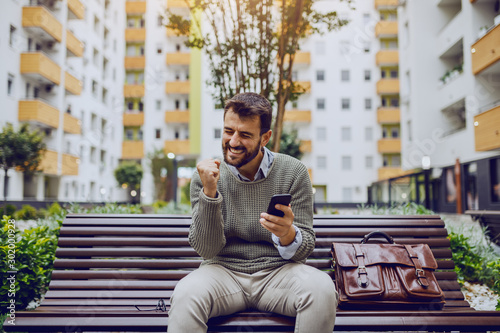 Successful handsome fashionable caucasian businessman sitting on bench in park, reading message on smart phone and rejoicing for achievement. Next to him is leather handbag. photo