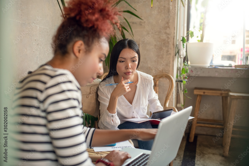 Dark-haired businesswoman looking at laptop sitting near friend Dark-haired businesswoman looking at laptop sitting near friend