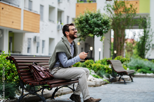 Side view of laughing caucasian stylish businessman sitting on bench after work and using smart phone. Next to him is handbag.