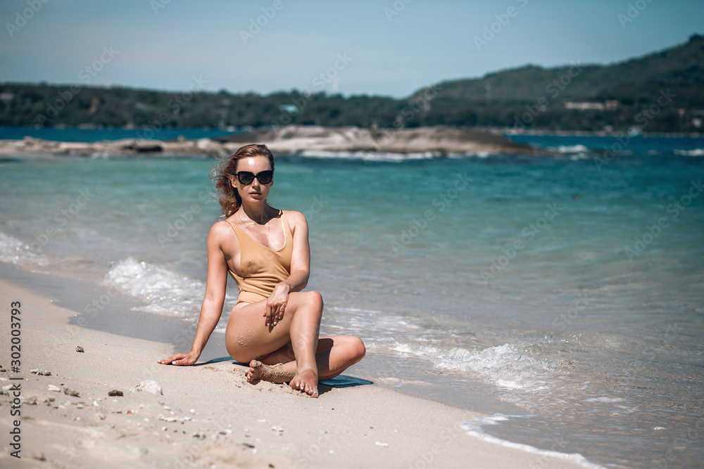 An amazing woman sitting on white sand in sexy beige swimsuit. A blondie with beautiful booty is alone on the beach with rocks. Kata beach, Phuket An amazing woman sitting on white sand in sexy beige swimsuit. A blondie with beautiful booty is alone on the beach with rocks. Kata beach, Phuket