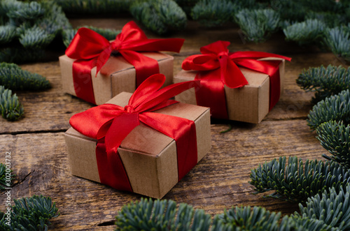 Close view of three gift boxes with red bows around which there are branches of fir on a wooden textured background.