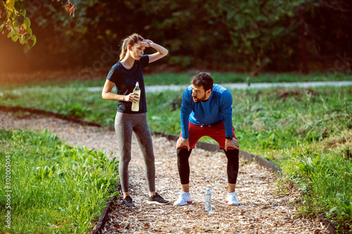 Tired caucasian sporty couple standing on trail in woods and resting from running. Woman holding bottle with refreshment. Both are in sportswear.