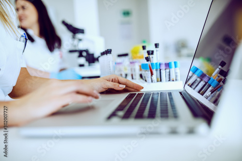 Close up of female lab assistant in white uniform sitting in lab and using laptop for data entry Fototapet