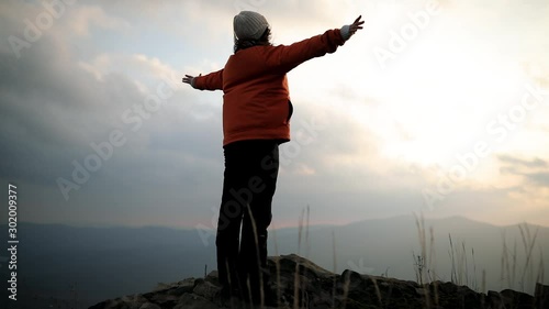 The camera revolves around a young beautiful woman standing on the edge of a cliff at the top of a mountain and raises her hands up to meet the sun. Happy and drunk on life, youth and happiness
