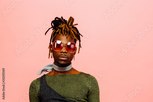Portrait of a serious young man in green with dreadlocks and red sunglasses, isolated on pink