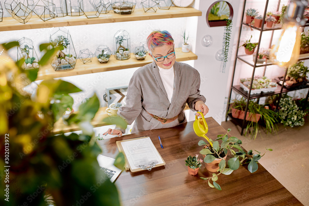 Beautiful elderly business lady wearing modern blazer, eyeglasses, with short colorful hair carefully watering plants in her modern full of green office
