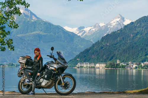 Pretty women is standing with traveler motorcycle. Vacation and hobby concept, jorney on two wheels. Sunny summer day in the Alpine mountains. Zell am see lake on background Austria. Copy space