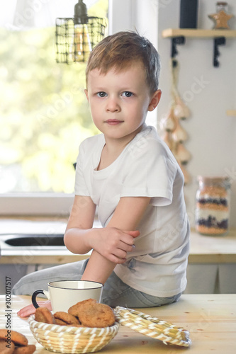 Cute little boy portrait in a white t-shirt sitting on wooden table. Vitamine breakfast in bright kitchen room interior.