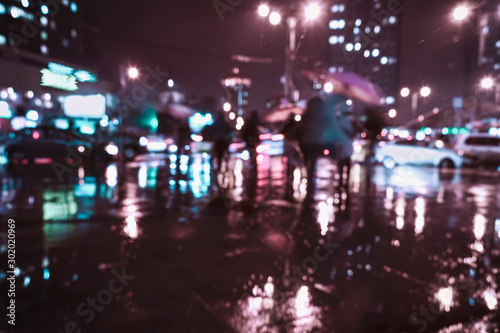 Blurred modern city at rayny night, defocused urban bokeh city lights, silhouettes of the walking people under umbrellas. Abstract background © svetlanais