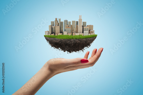 Side closeup of woman's hand facing up and levitating small piece of land with modern city on it on light blue background.