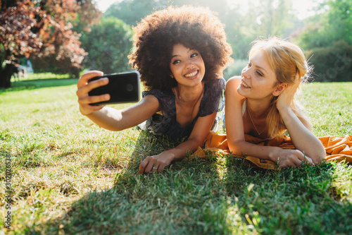 Couple of young women take a selfie with the smart phone lying on the grass of a meadow on a summer day at the park - Millennials have fun together laughing and joking