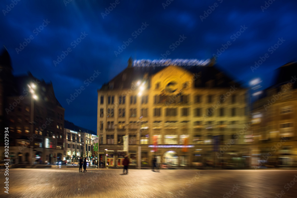 Blurry motion image of people walking on Dam square in Amsterdam. It is a rainy summer night with cloudy, dark blue color sky.