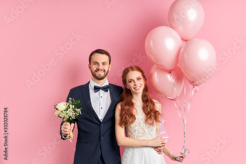 Handsome man in tuxedo and redhaired woman in wedding dress look at each other with love. Holding pink air balloons and bouquet, pink background