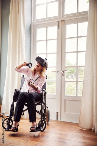 Mature Disabled Woman In Wheelchair At Home Wearing Virtual Reality Headset With Gaming Controllers © Monkey Business