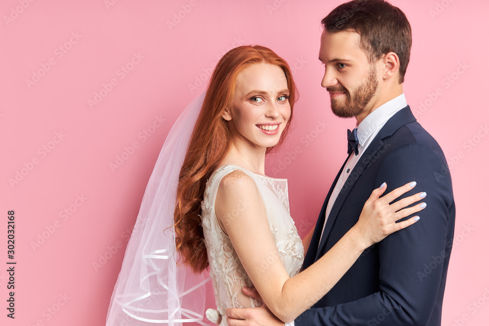 Caucasian man in tuxedo and woman in white wedding dress lovely hug each other and smile, after wedding ceremony posing isolated over pink background. Caucasian man in tuxedo and woman in white wedding dress lovely hug each other and smile, after wedding ceremony posing isolated over pink background.