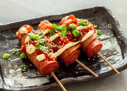 Bacon wrapped hot dogs on a stick. Topped with mayo, ketchup, spring onion, sesame, and spring onion. photo