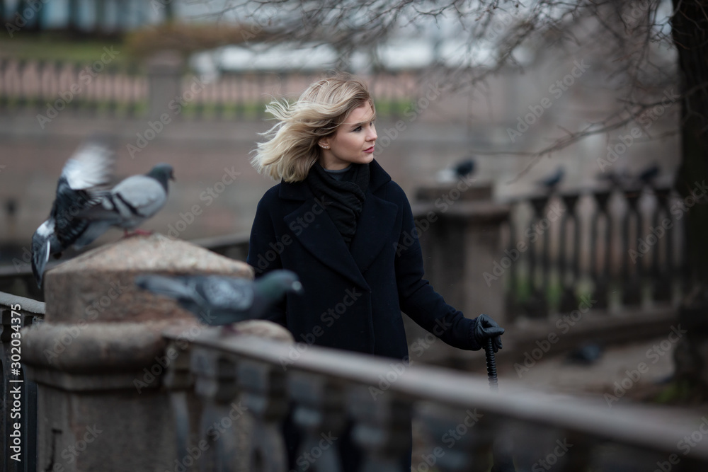 Beautiful young woman in a coat walks along the fence on the river embankment. Attractive girl with blond hair fluttering in the wind. Cool autumn weather. Lifestyle, walks around the city and travel.