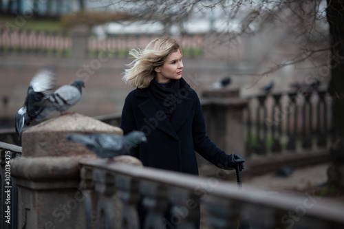 Beautiful young woman in a coat walks along the fence on the river embankment. Attractive girl with blond hair fluttering in the wind. Cool autumn weather. Lifestyle, walks around the city and travel.
