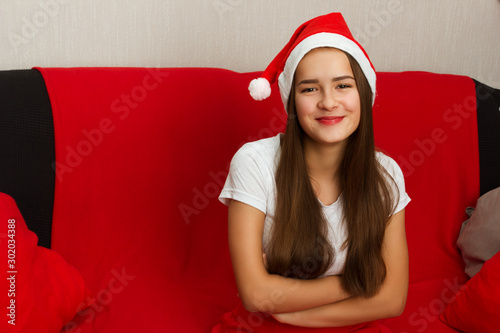 Christmas mood. A young European girl with long dark hair in a Santa Claus hat is sitting on the sofa and smiling.