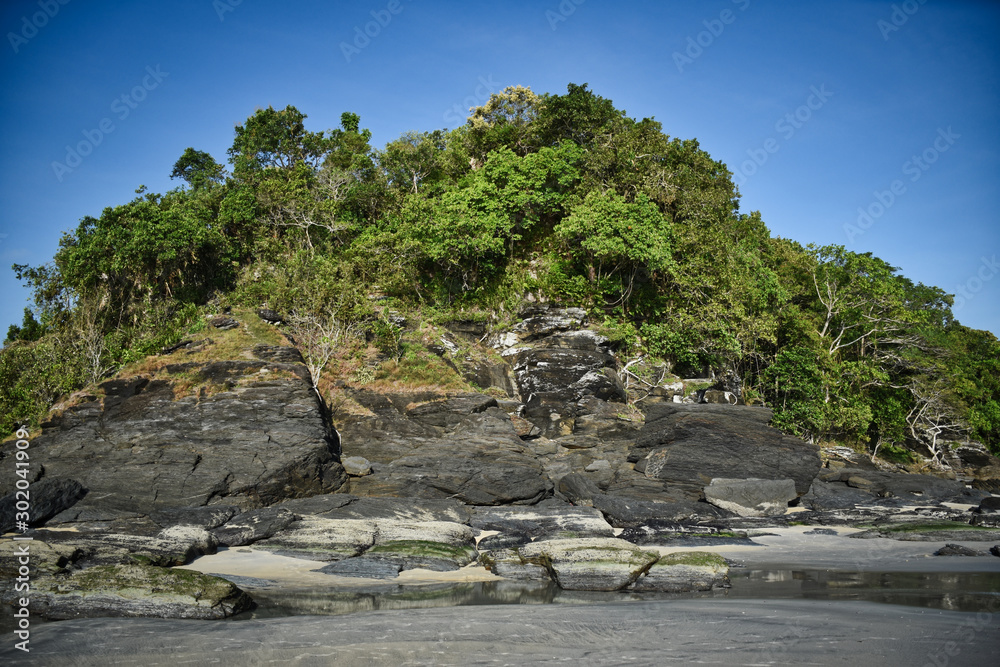 Hillside with black rocks and green trees, plants on the shores of the sandy beautiful exotic and stunning Cenang beach in Langkawi island
