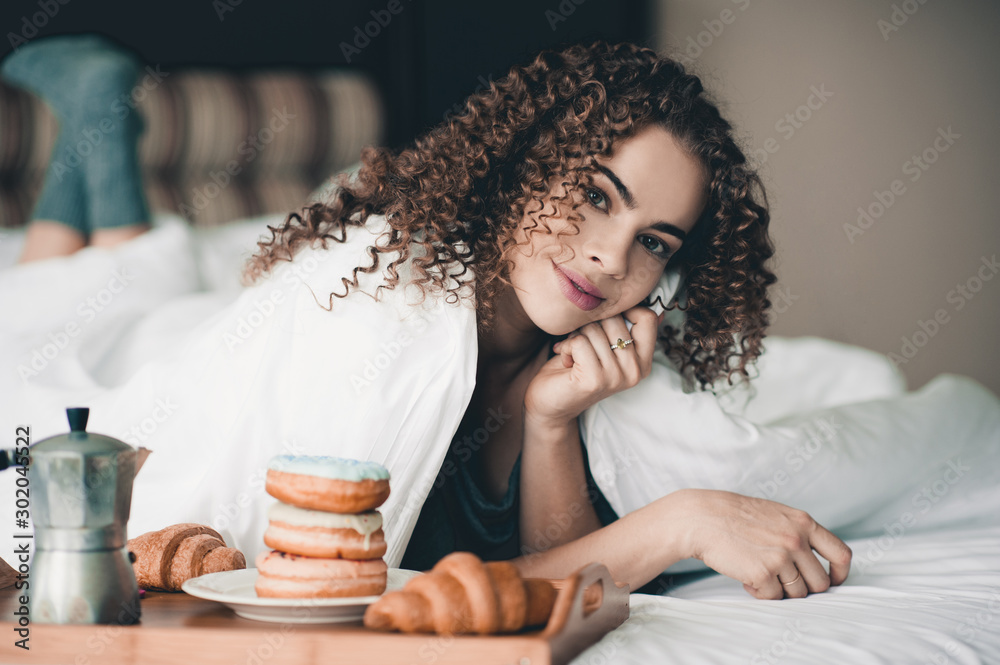 Cute woman wake up with breakfast on wooden tray: stack of donuts, coffee pot and croissant lying in bed closeup. Good morning. Looking at camera. Breakfast time. - obrazy, fototapety, plakaty 