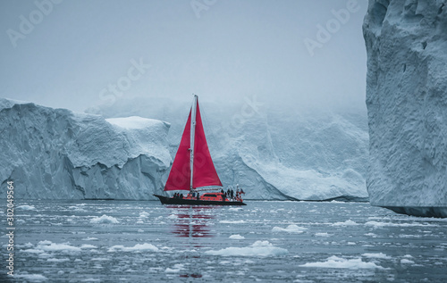 A small boat among icebergs. Sailboat cruising among floating icebergs in Disko Bay glacier during midnight sun Ilulissat, Greenland. Studying of a phenomenon of global warming Ices and icebergs