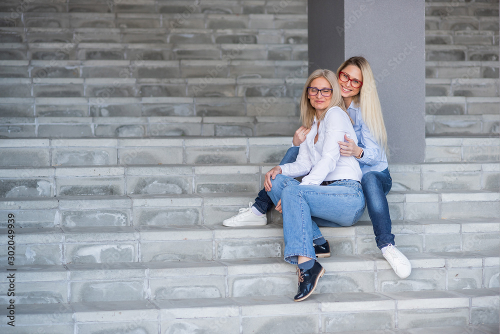 A beautiful blonde with glasses and her mature mother are sitting on the stairs outdoor. A well-groomed elderly woman hugs her adult daughter tightly. Love and family values. Mothers Day. A beautiful blonde with glasses and her mature mother are sitting on the stairs outdoor. A well-groomed elderly woman hugs her adult daughter tightly. Love and family values. Mothers Day.