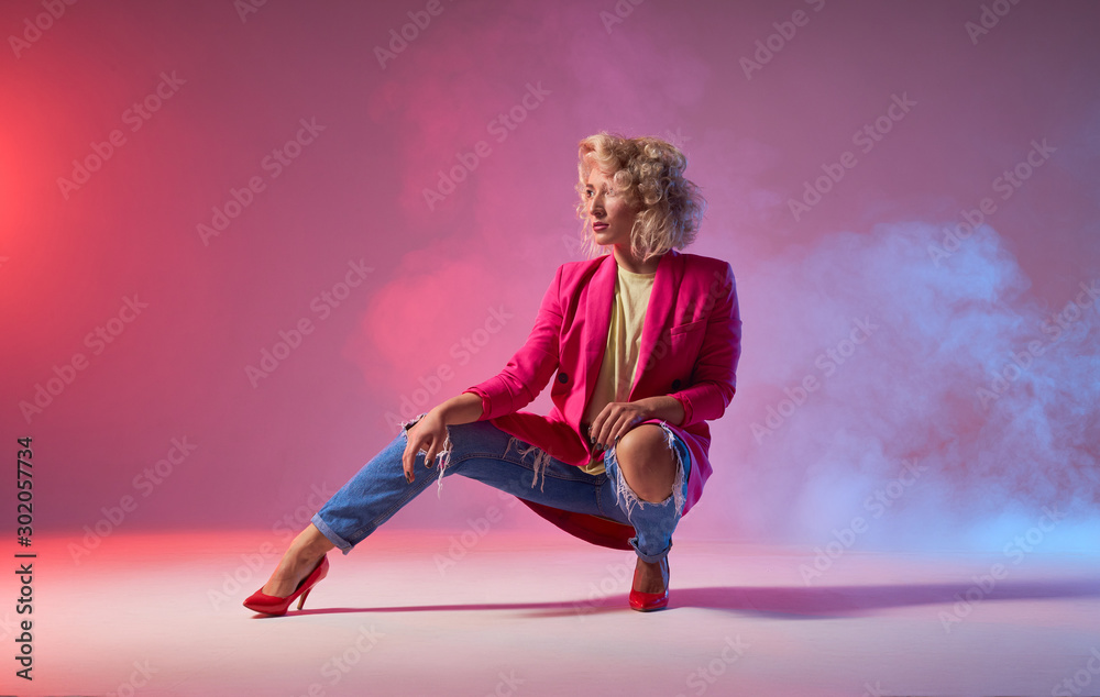 Slim tanned woman dressed in stylish clothes, and red heels, sitting on squat, looking away, showing dancing element on studio background, jazz funk street hip hop music