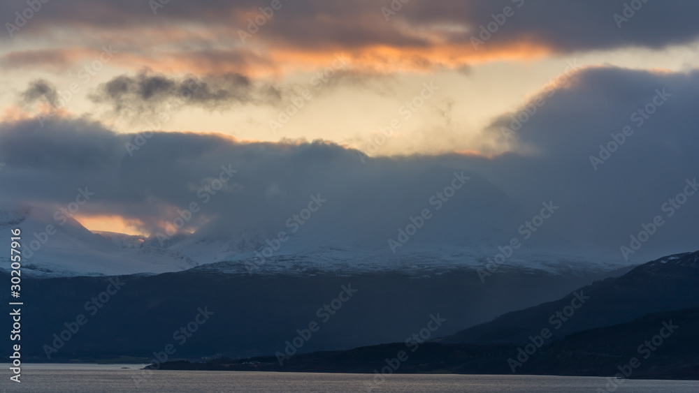 Mountains by a fjord with some snow and dark clouds in sunset colored sky in northern Norway