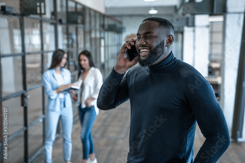 Quick business talk. Handsome young african american smiling man talking on the phone and gesturing while his colleagues standing in the background