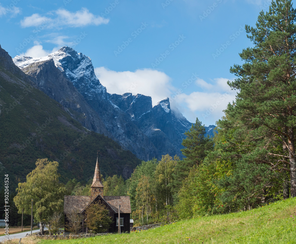 Kors Church, old wooden parish church in Rauma in Romsdal valley, Norway with road E136, green forest and mountain massif Trolltindene, Troll wall Trollveggen. Summer blue sky white clouds.