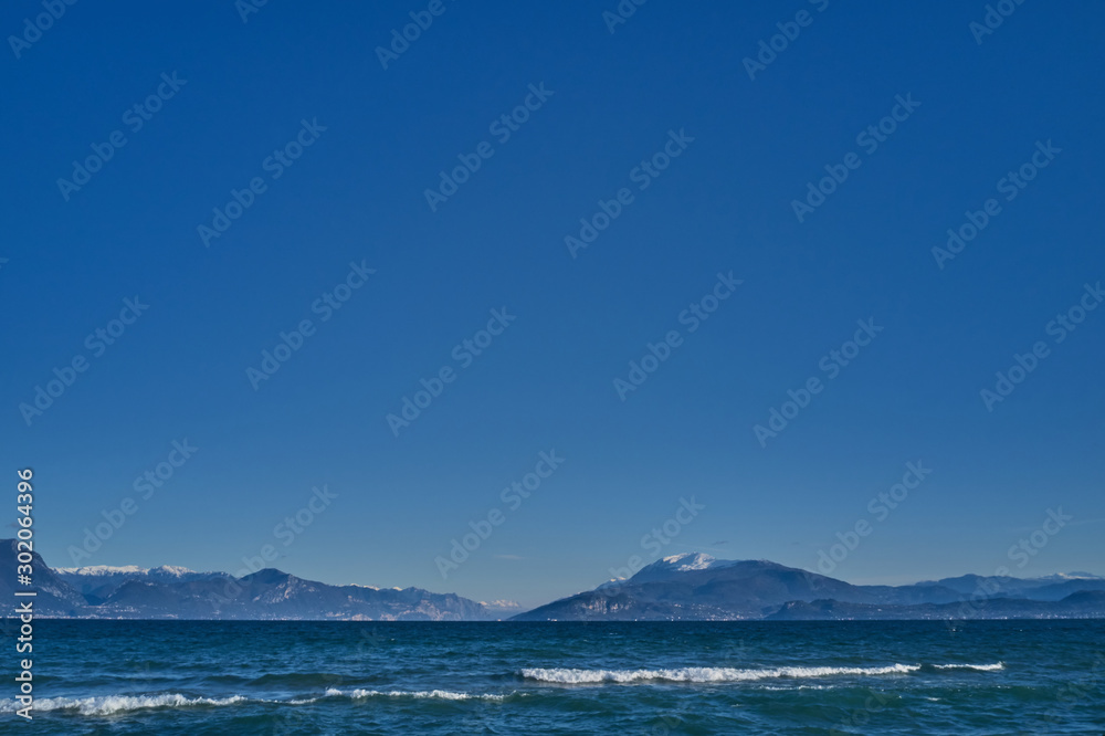 Panoramic view of Lake Garda the north of Italy. Mountains in the snow. White wave crest on a background of mountains and blue sky Panoramic view of Lake Garda the north of Italy. Mountains in the snow. White wave crest on a background of mountains and blue sky