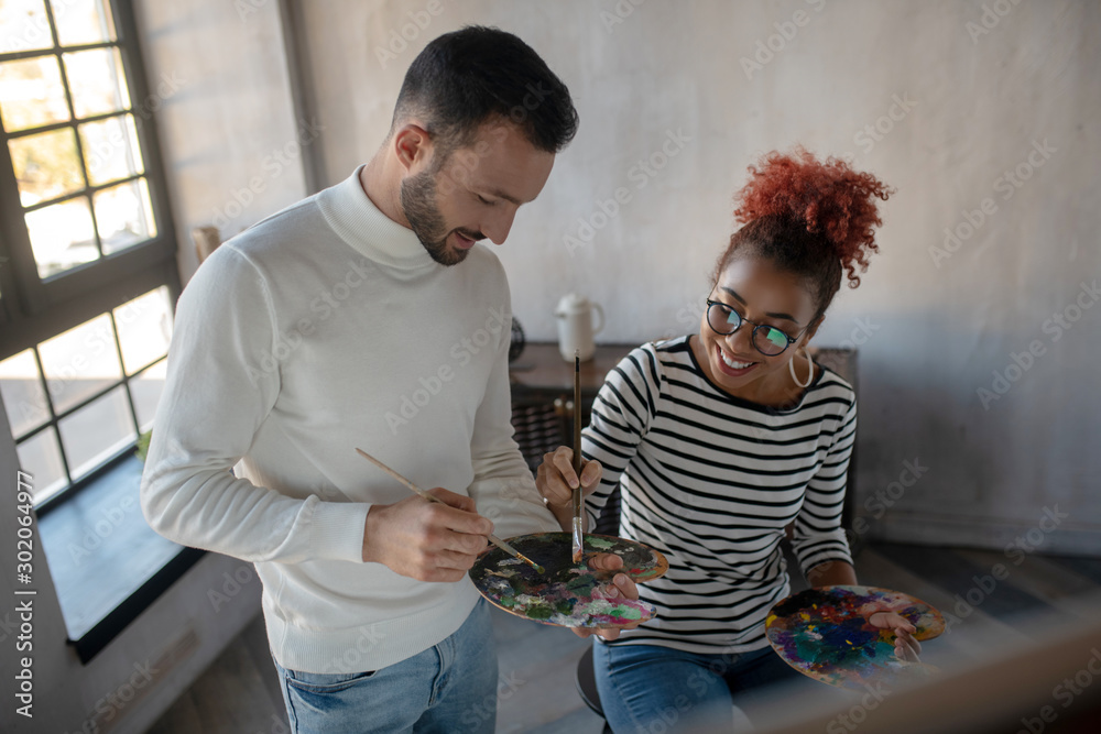 Fototapeta premium Smiling girlfriend taking paints while painting with beloved