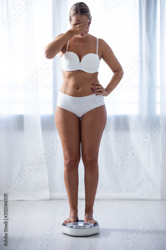 Worried young woman covering her eyes while weighing herself on the weight scale at home.