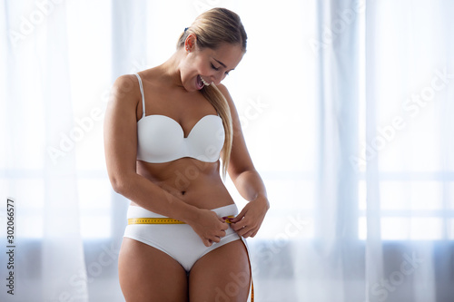 Fototapeta Happy young woman because she has lost weight measuring her body at home