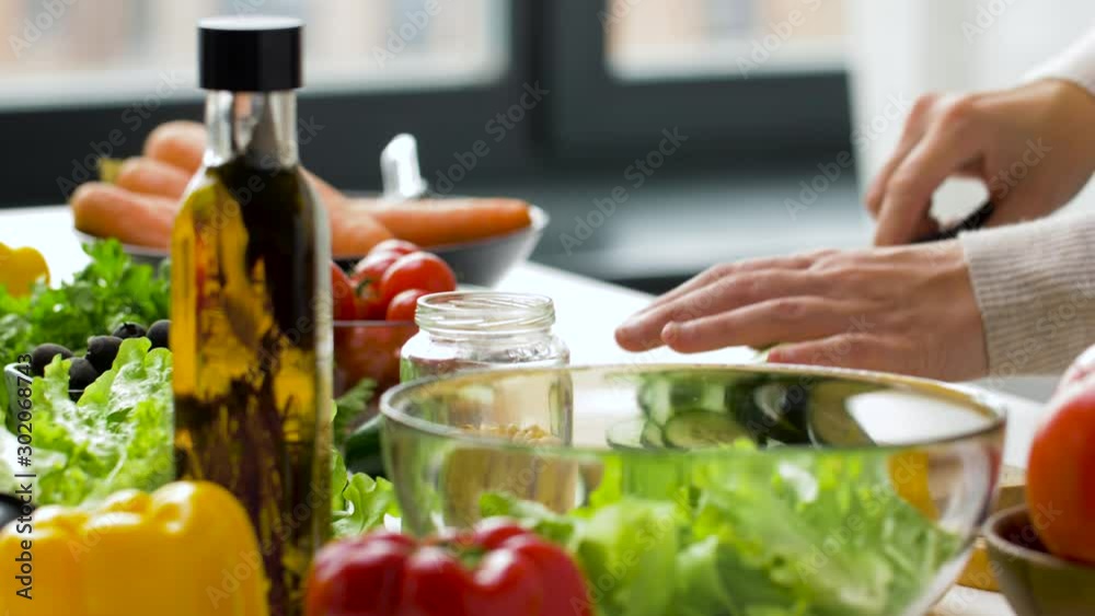 healthy eating, vegetarian food and cooking concept - young woman making vegetable salad and chopping cucumber with kitchen knife on wooden cutting board at home