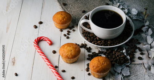 A hot cup of coffee with the coffee beans, small cupcakes and a candy on the table. Composition of a delicious coffee and sweet snacks.