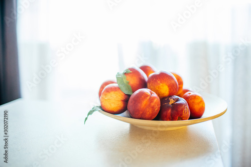 Fototapeta Naklejka Na Ścianę i Meble -  Fresh ripe peaches with leaves on a wooden plate on a kitchen table against the window. Organic food concept and prevention of vitamin deficiency. Fruits for cooking.