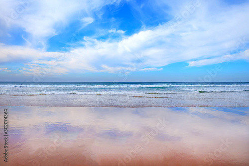 big blue and white sky a blue sea and waves and clear wet sandy beach reflecting the sky