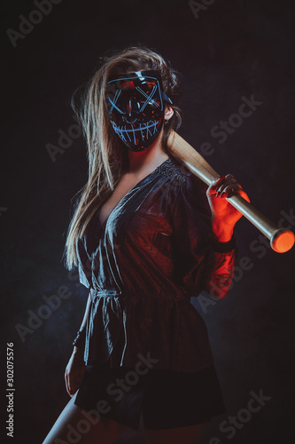 Lawbreaker female in spooky mask is standing at dark studio with bat in her hands.