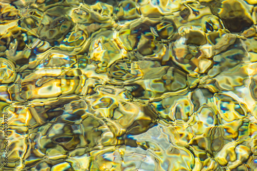 Abstract background of clear sea or river water ripple waves in light green khaki color. Sea water with different color pebbles at the bottom. Sun reflection on sea water surface.