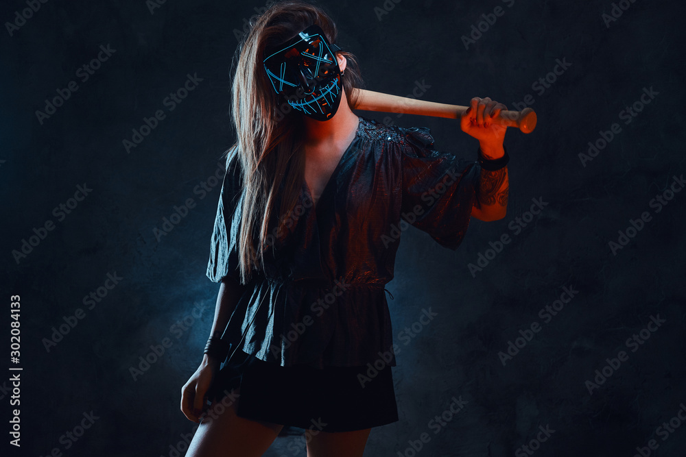 Lawbreaker female in spooky mask is standing at dark studio with bat in her hands. Lawbreaker female in spooky mask is standing at dark studio with bat in her hands.