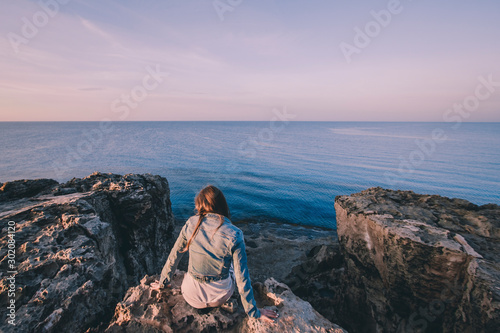 Woman traveler sitting near sea on top of cliff in summer mountains and enjoying view of sea and nature. Cape Greco, Cyprus, Mediterranean Sea. Sunrise