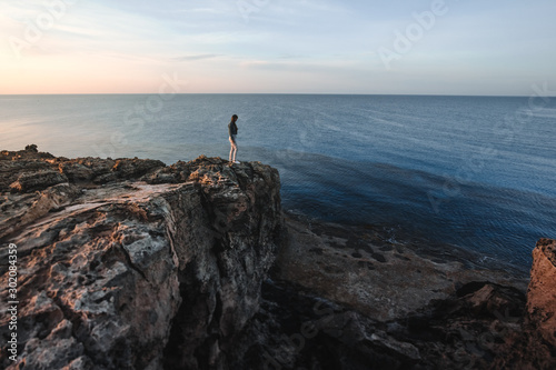 Woman traveler standing near sea on top of cliff in summer mountains and enjoying view of sea and nature. Cape Greco, Cyprus, Mediterranean Sea. Sunrise