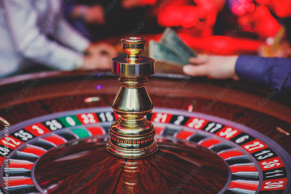A close-up vibrant image of multicolored casino table with roulette in motion, with casino chips. the hand of croupier, mone and a group of gambling rich wealthy people in the background A close-up vibrant image of multicolored casino table with roulette in motion, with casino chips. the hand of croupier, mone and a group of gambling rich wealthy people in the background