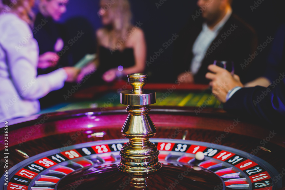 A close-up vibrant image of multicolored casino table with roulette in motion, with casino chips. the hand of croupier, mone and a group of gambling rich wealthy people in the background A close-up vibrant image of multicolored casino table with roulette in motion, with casino chips. the hand of croupier, mone and a group of gambling rich wealthy people in the background